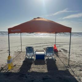 Beach scene with orange canopy, two chairs, cooler, and sunlight.