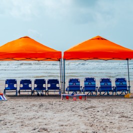 Two orange beach canopies with blue chairs on a sandy shore.